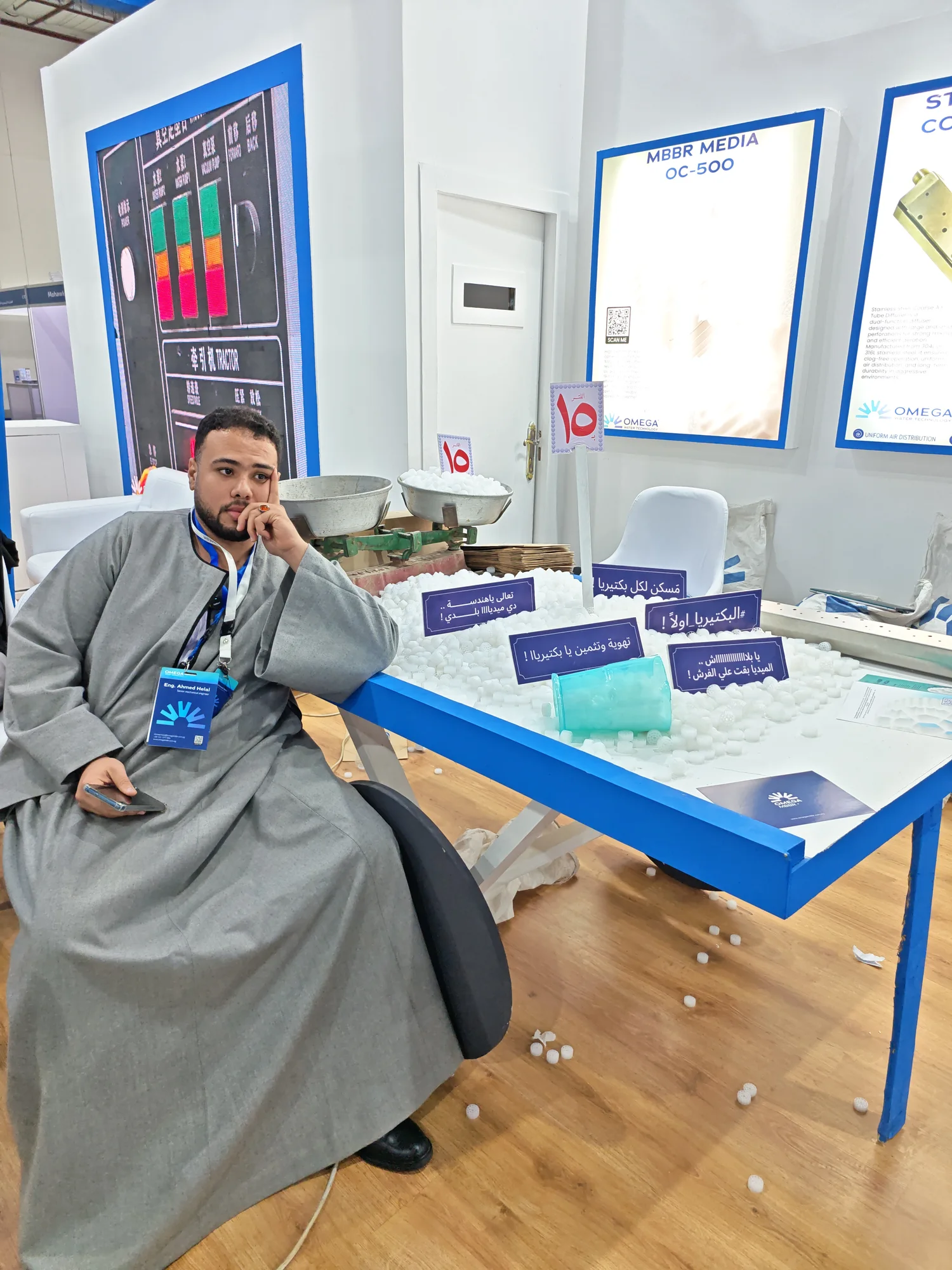 أحمد هلال يجلس بجوار عربية محملة بميديا MBBR وميزان كفتين في معرض أوميجا